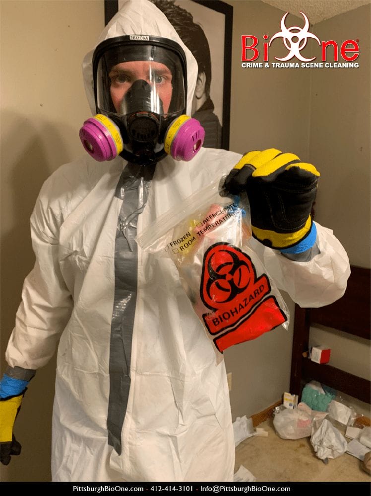 Image shows Bio-One technician dressed in hazmat and PPE holding a small red bag of biohazardous waste.